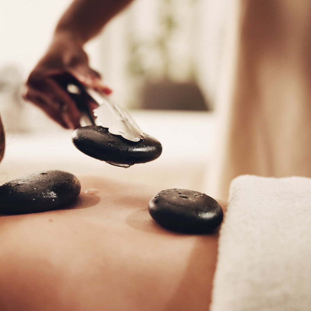Hot stone massage with smooth black stones placed on a person's back, while a therapist holds one stone. A white towel covers part of the body, and the scene is softly lit in warm spa tones.