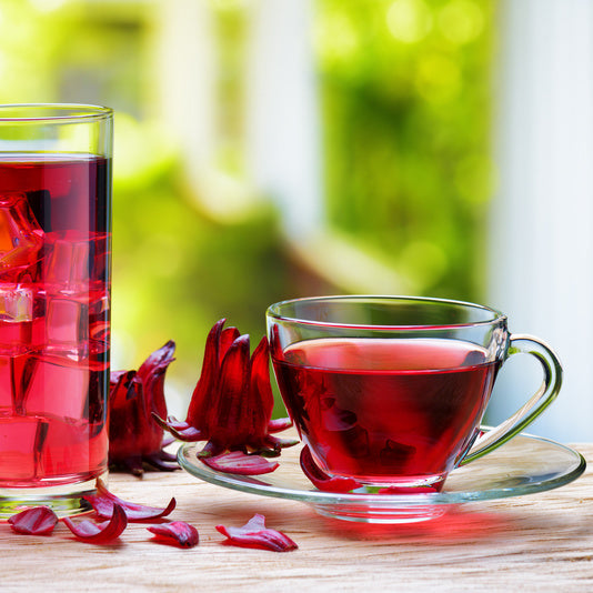 Clear glass cup and glass filled with red dried hibiscus tea on a wooden surface with blurred greenery in the background.