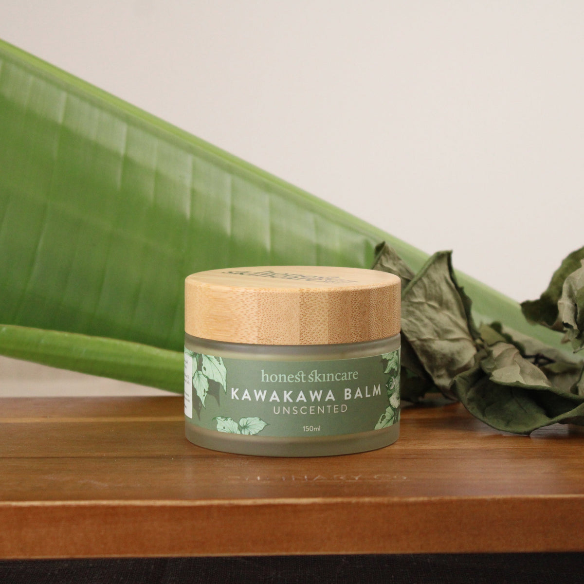Kawakawa balm in a jar with bamboo lid, surrounded by green leaves on a wooden surface.