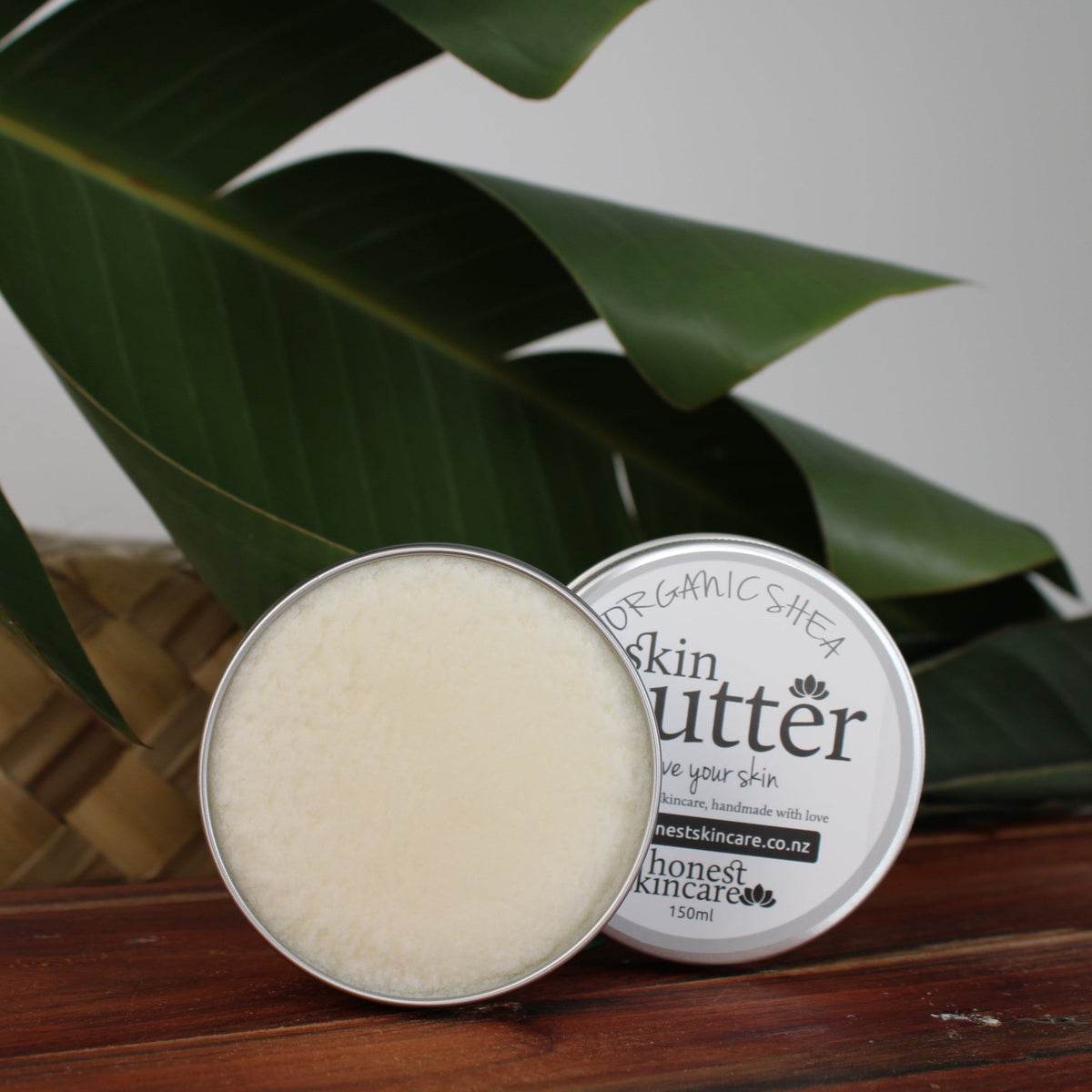 Shea butter body cream in a tin on a wooden surface surrounded by tropical leaves.