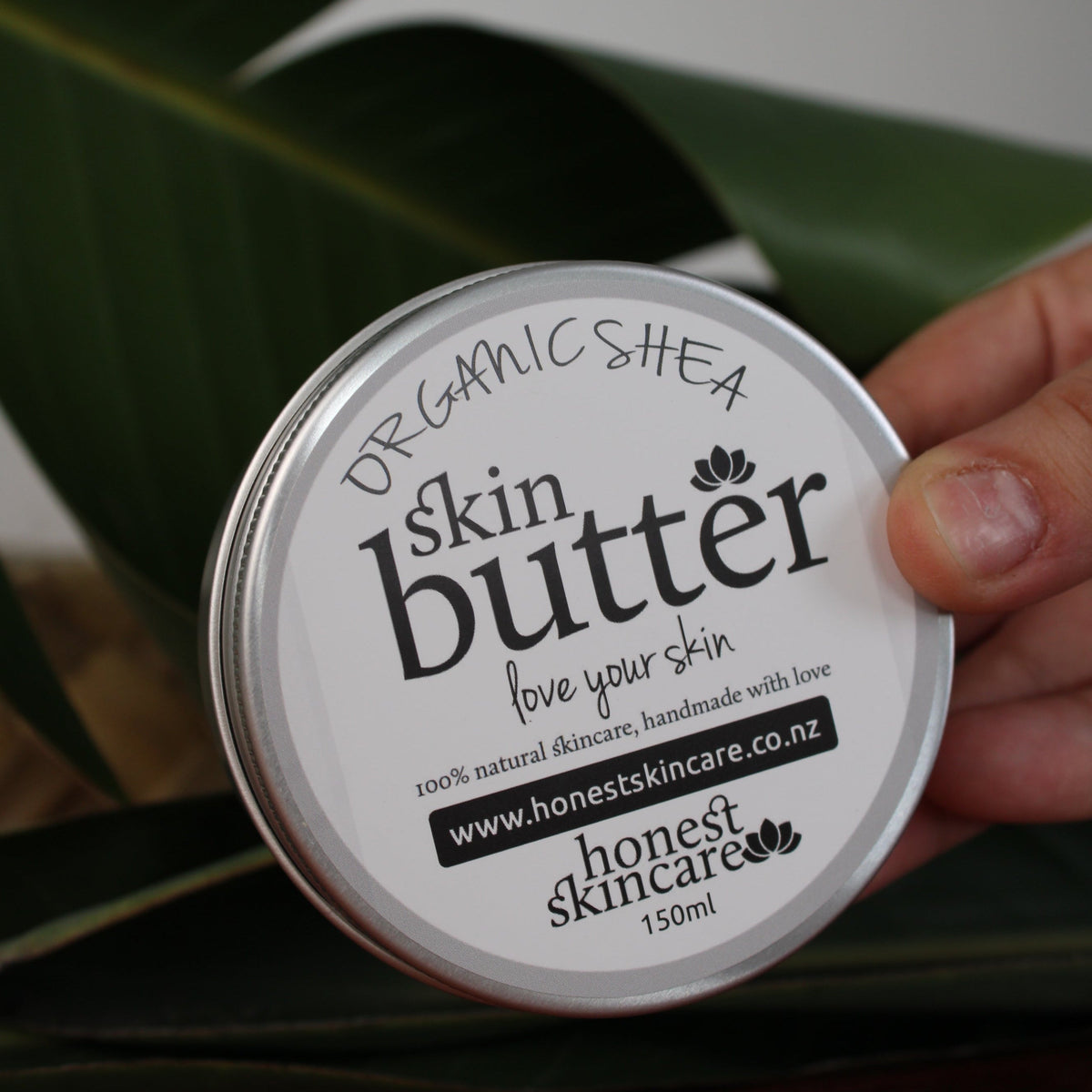 Shea butter body cream displayed in a tin with a hand, surrounded by tropical leaves.